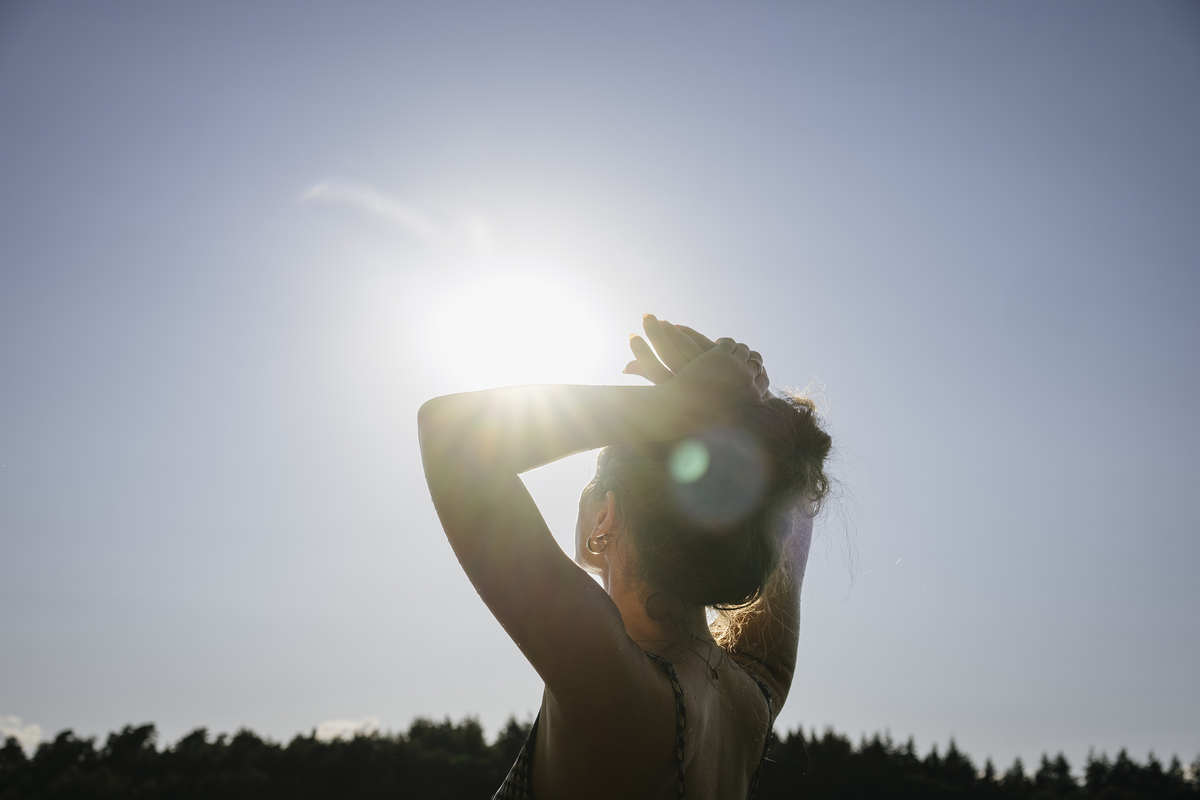 Frau steht im Gegenlicht der Sonne, hält ihre Haare hoch und blickt in den hellen Himmel