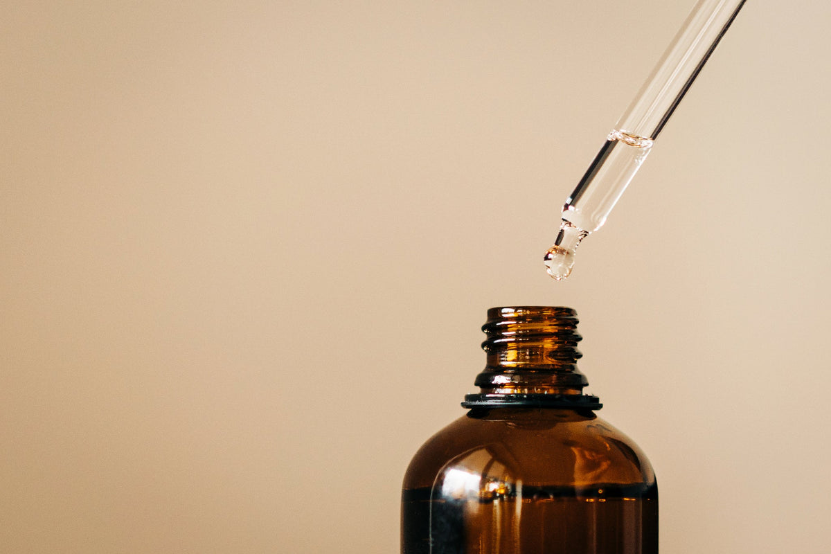 Braune Glasflasche mit Pipette, aus der ein Tropfen Öl fällt, vor neutralem Hintergrund