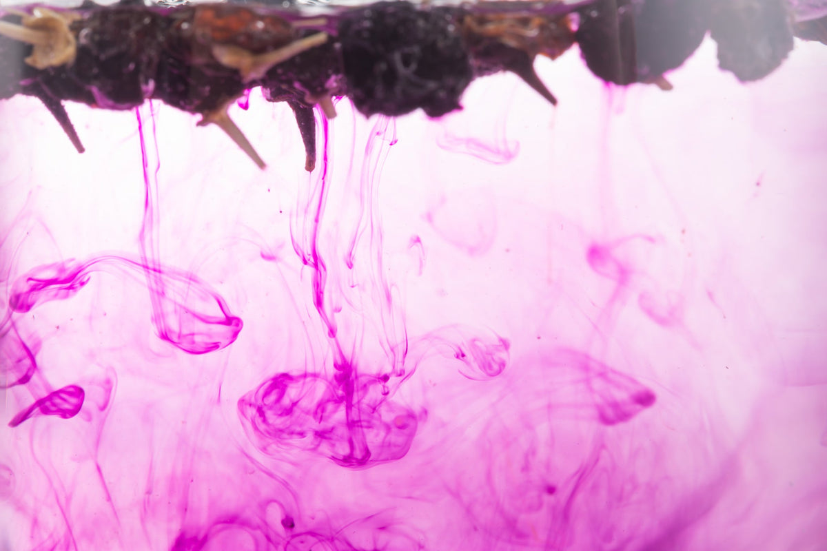 Glasflasche mit rosa Flüssigkeit, daneben geöffnete Granatapfelfrucht und lose Granatapfelkerne auf einem Holztisch.