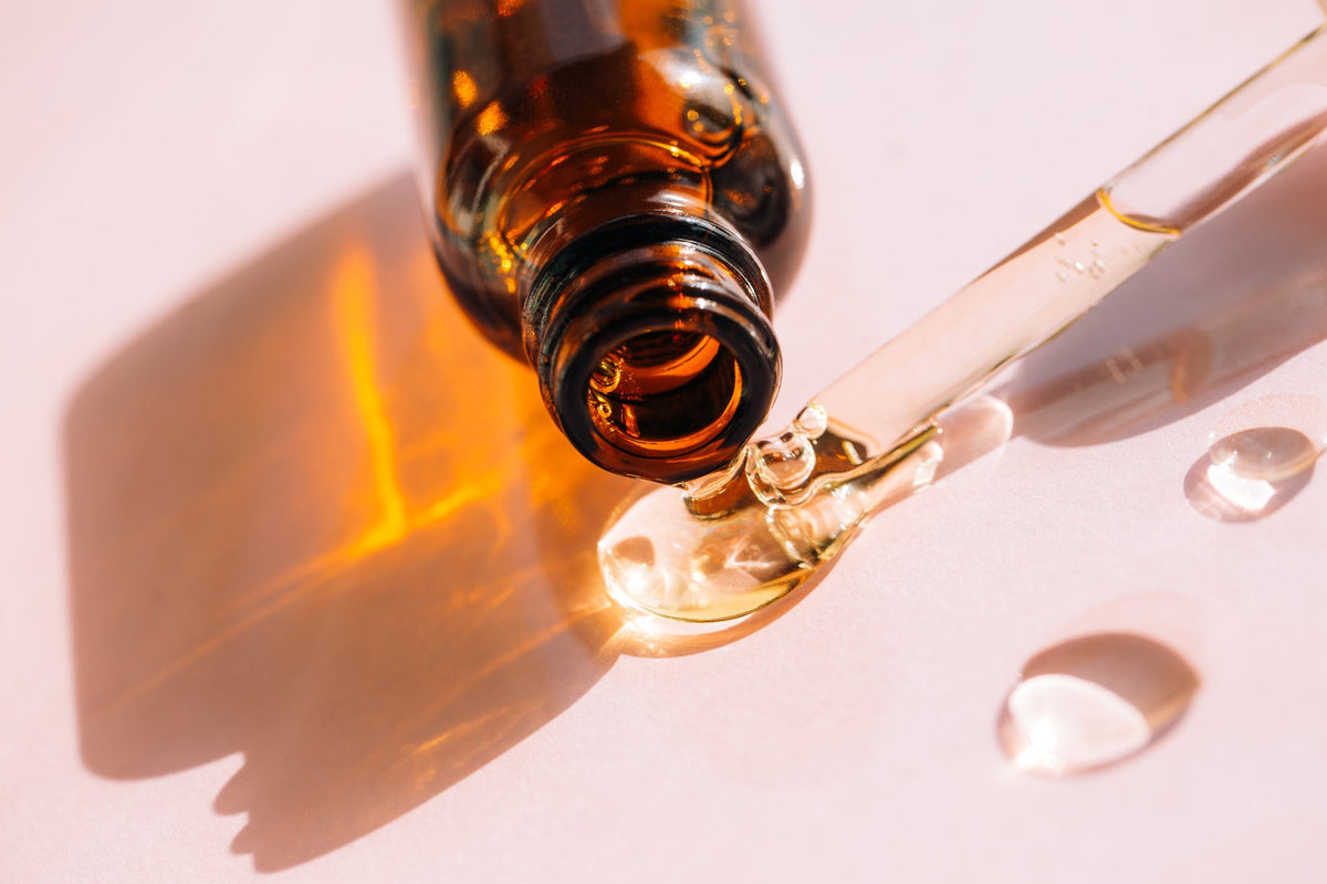 Braunglasflasche mit Pipette, aus der Hyaluron-Serum auf rosafarbenem Hintergrund tropft.